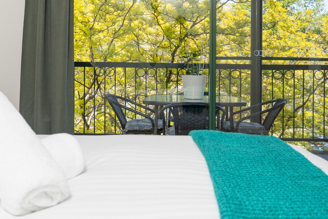 Une chambre d’hôtel avec un lit, un plaid vert, une table à manger avec des chaises et une vue sur les arbres, visible à travers la rambarde du balcon.