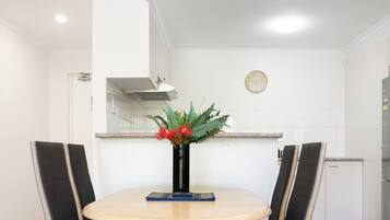 A dining area with a round table, two chairs, a vase with flowers, a clock on the wall, and a kitchen in the background.