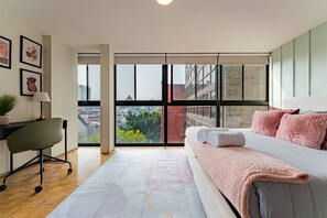A modern bedroom with a large bed, a desk, and a view of the city through floor-to-ceiling windows. - 7BR | 2 Modern Apartments Near Reforma | Sleeps 14 (Mexico City)