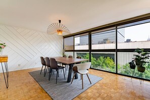 A modern dining area with a large table and chairs, a unique ceiling light, and a view of greenery through large windows. - 7BR | 2 Modern Apartments Near Reforma | Sleeps 14 (Mexico City)