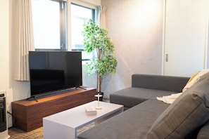 Ein modernes Wohnzimmer mit einem Flachbildfernseher auf einem Holzständer, einem grauen Sofa, einem weißen Couchtisch mit einem Buch und einer Topfpflanze.