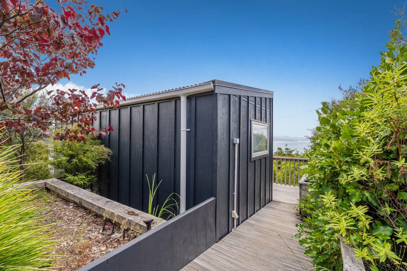 Un abri moderne, de couleur sombre, doté d’une petite fenêtre, entouré d’une terrasse en bois et d’une végétation luxuriante.