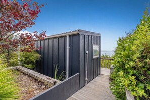 A modern, dark-colored shed with a small window, surrounded by a wooden deck and lush greenery.