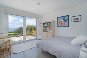 A bedroom with a large bed, a bookshelf, and a view through a sliding door to an outdoor deck. - Kuaotunu Heights | Epic Ocean Views & Outdoor Living (Kūaotunu)
