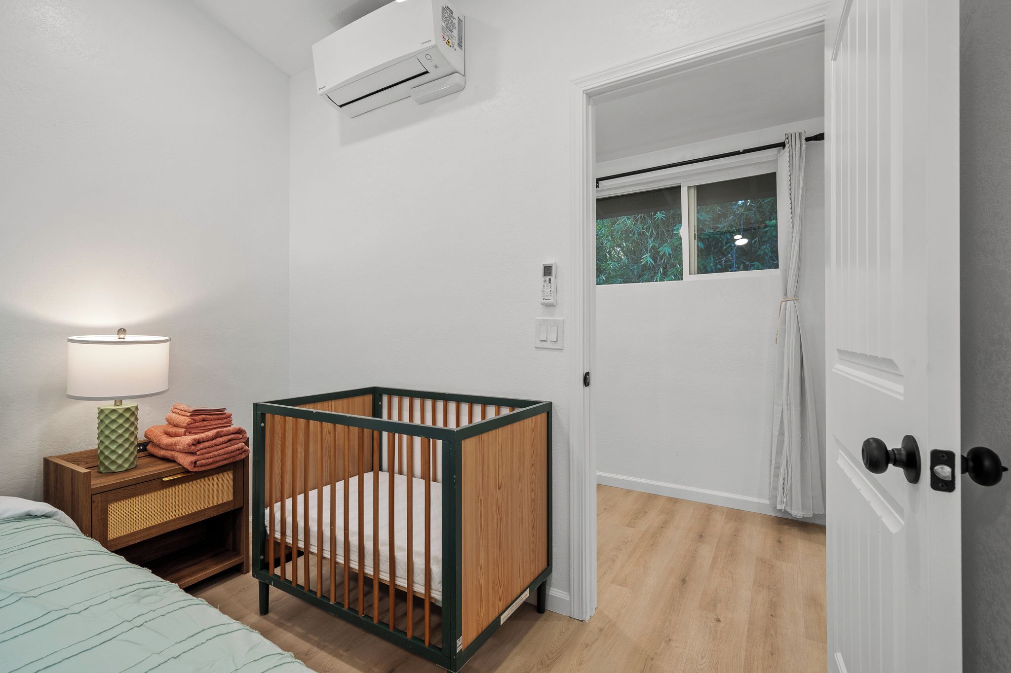 A room with a crib, a bedside table with a lamp, a wall-mounted air conditioner, and a door leading to another room.
