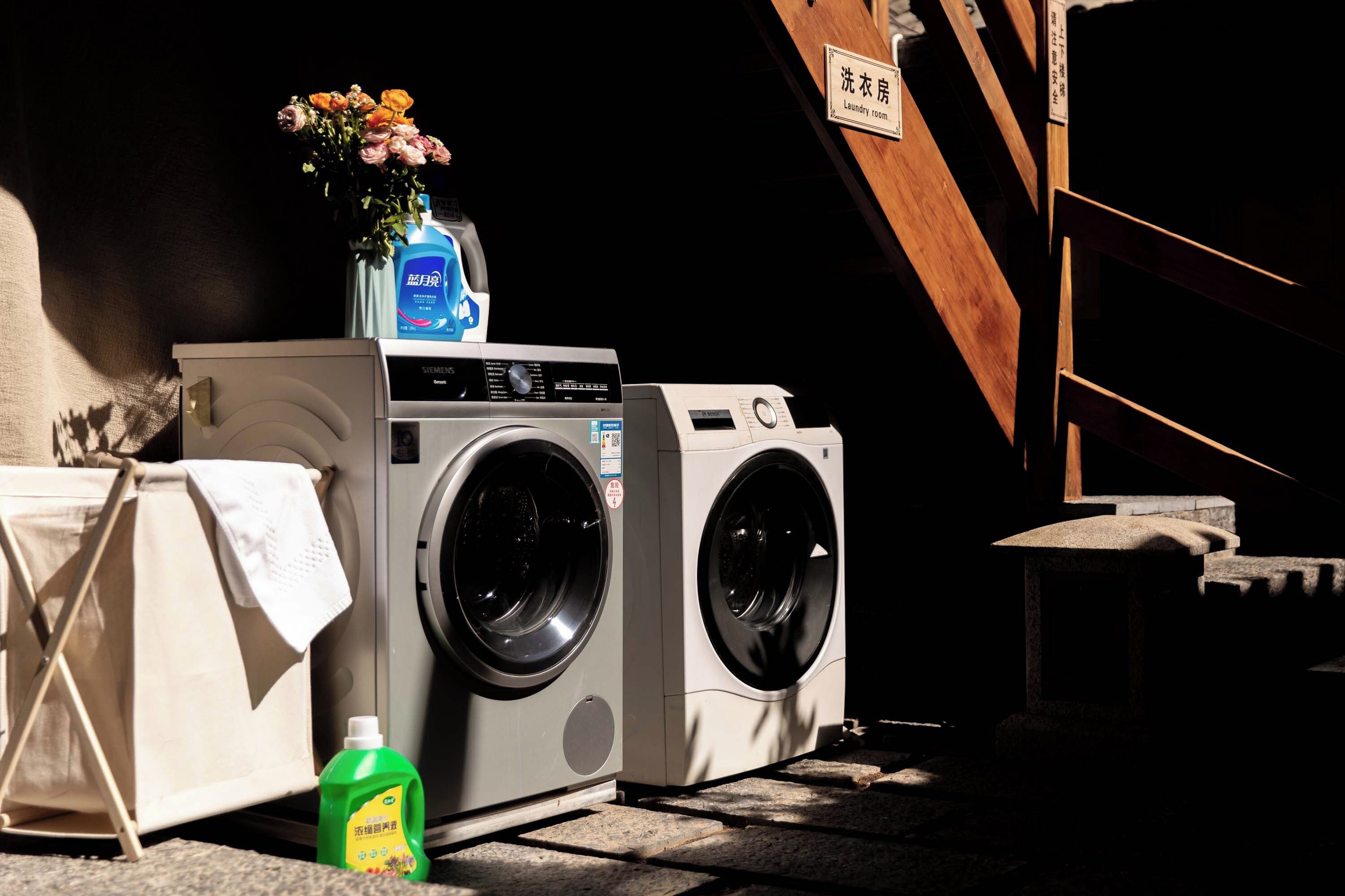 A laundry room with two washing machines, a vase with flowers, and a detergent bottle.
