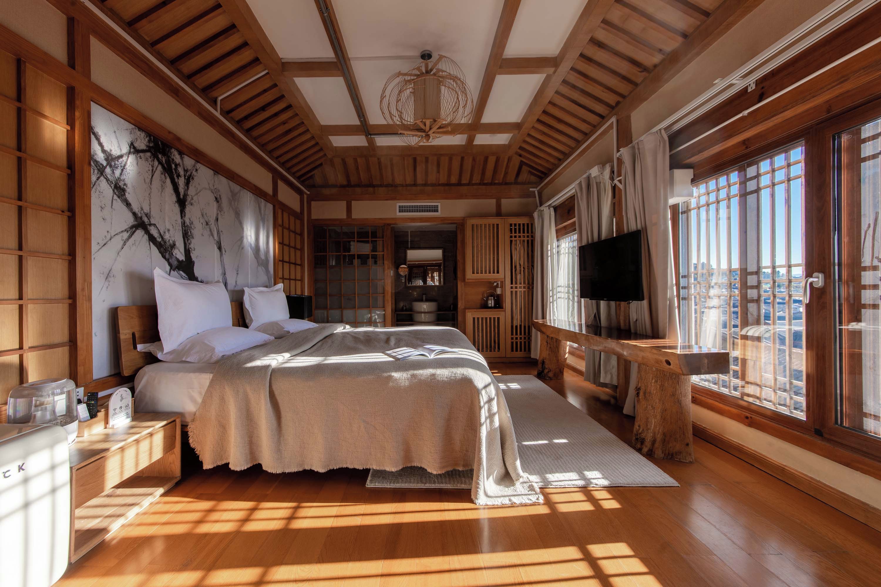 A traditional Japanese-style bedroom with tatami flooring, a large bed with white bedding, a television, and a view of the city through large windows.