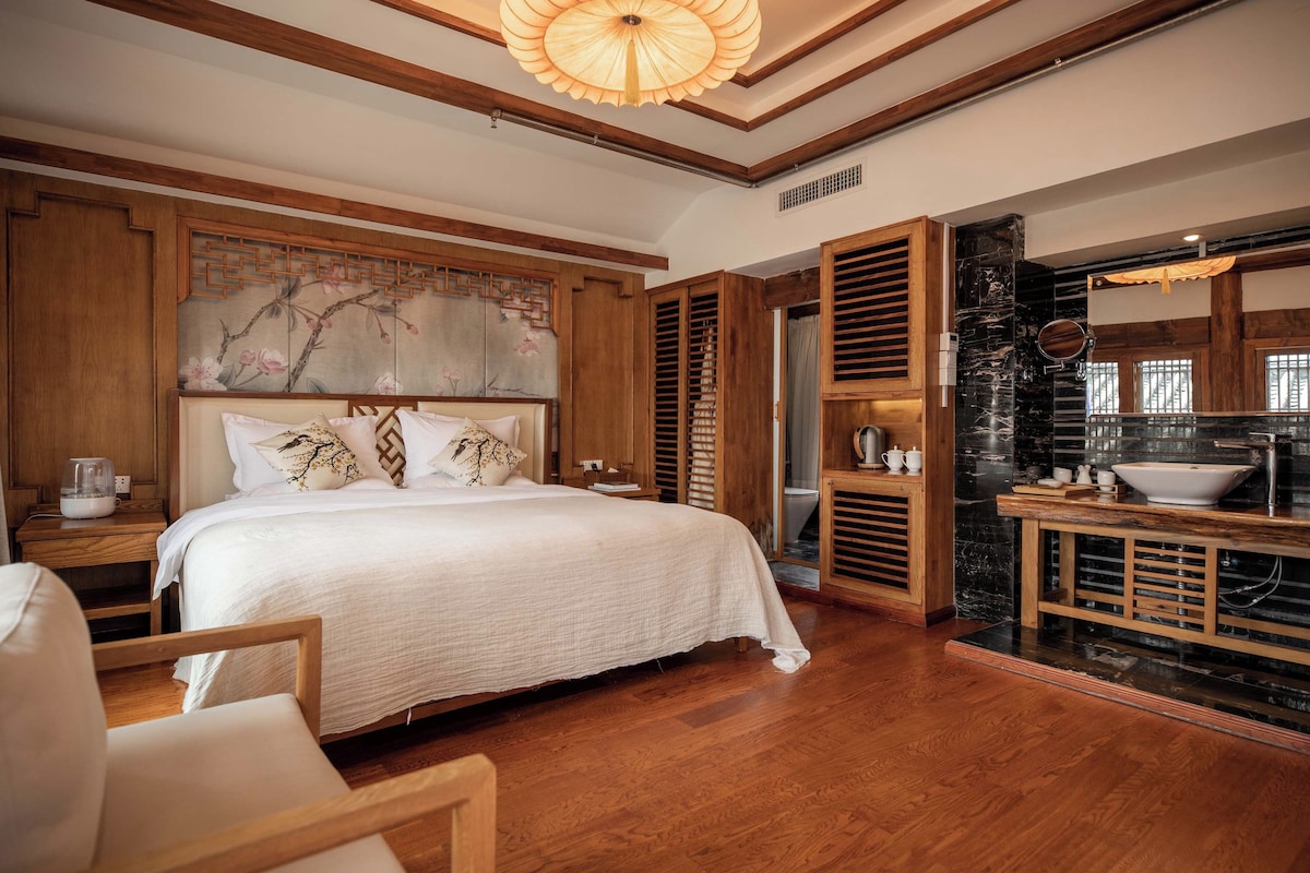 Quarto de hotel com uma cama grande, painéis de madeira, uma pintura e banheiro com pia e espelho.