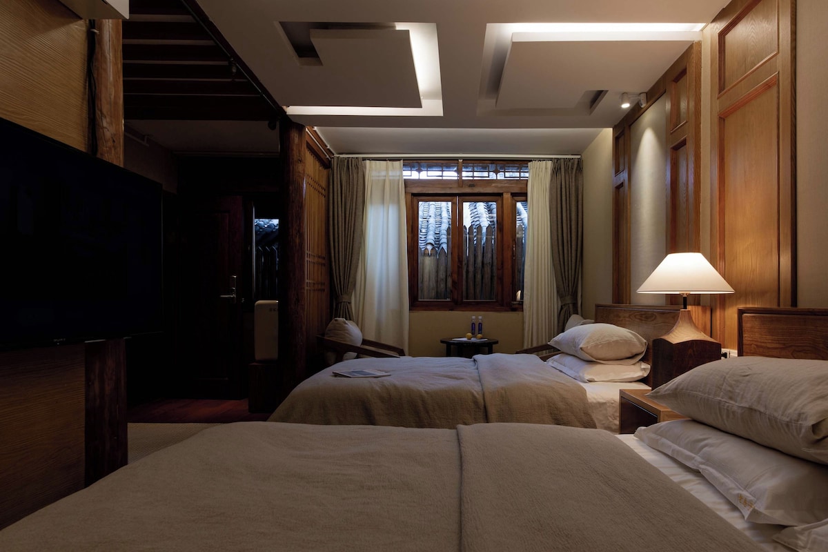A hotel room with two beds, a television, a window with curtains, and a lamp.