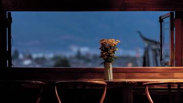 Una mesa de madera con un florero de flores frente a una ventana con un persiano abierto.