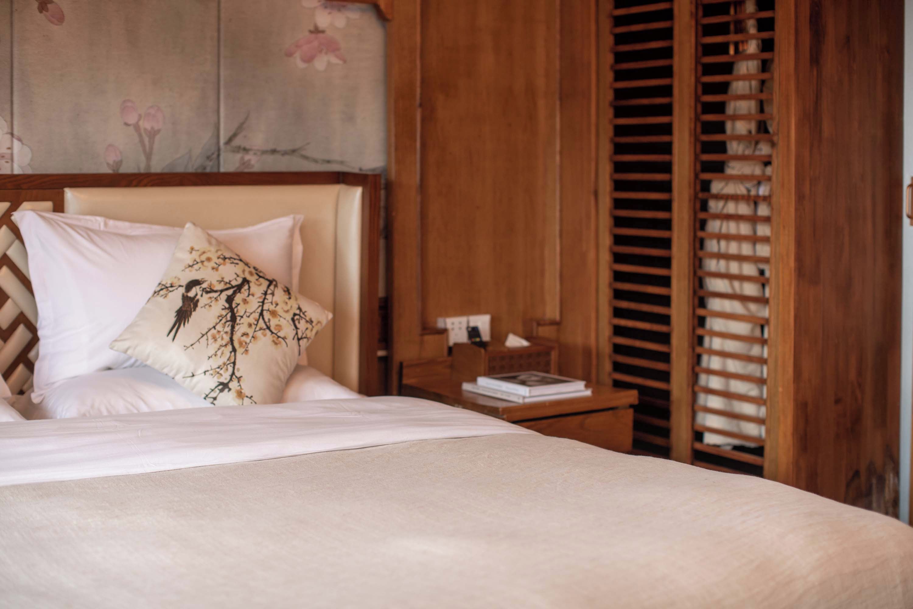 A neatly made bed with white linens and a decorative pillow, a wooden nightstand with a book and a lamp, and a wooden wardrobe with a sliding door.