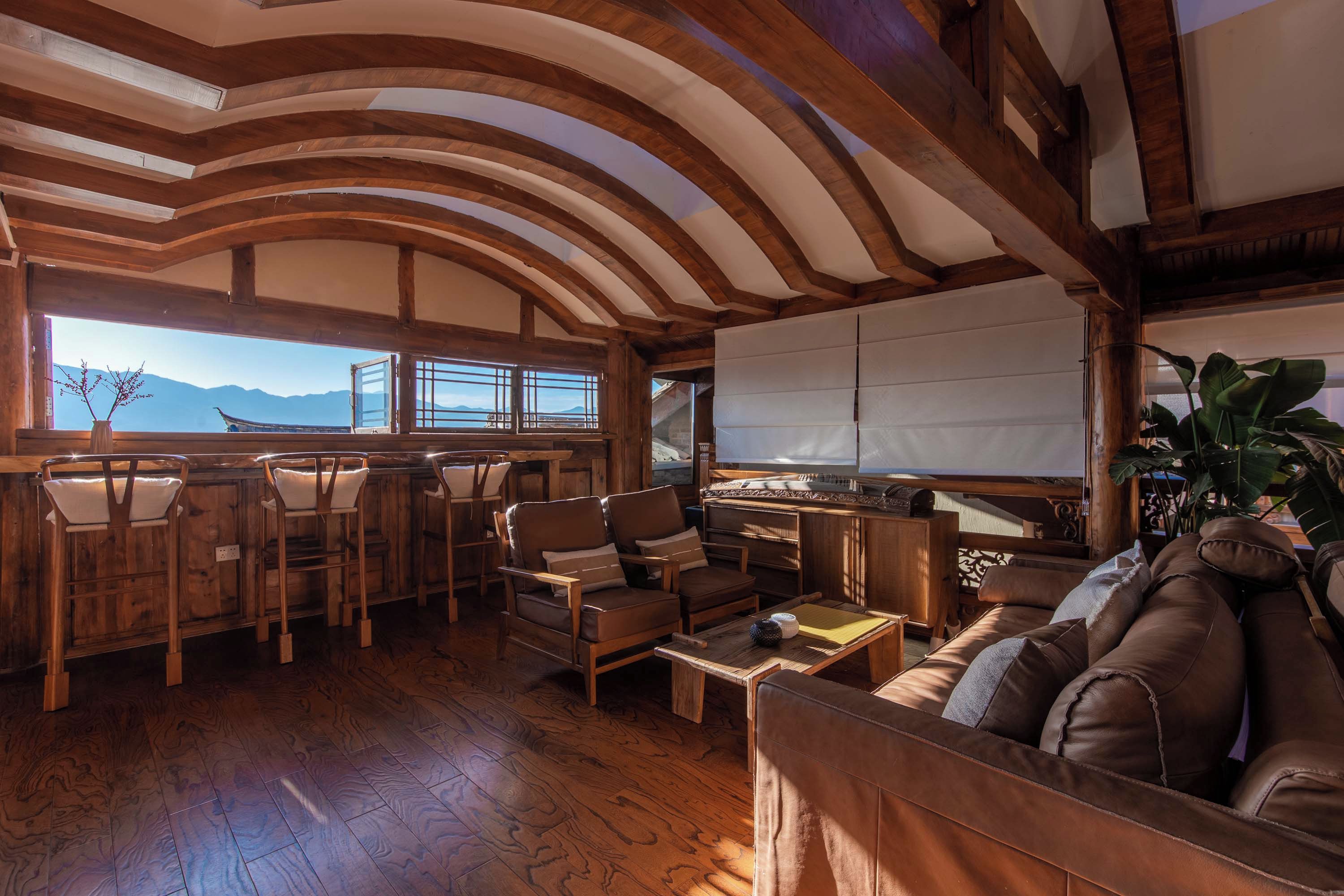 A wooden interior with a high ceiling, a seating area with chairs and a sofa, and a view of mountains through large windows.