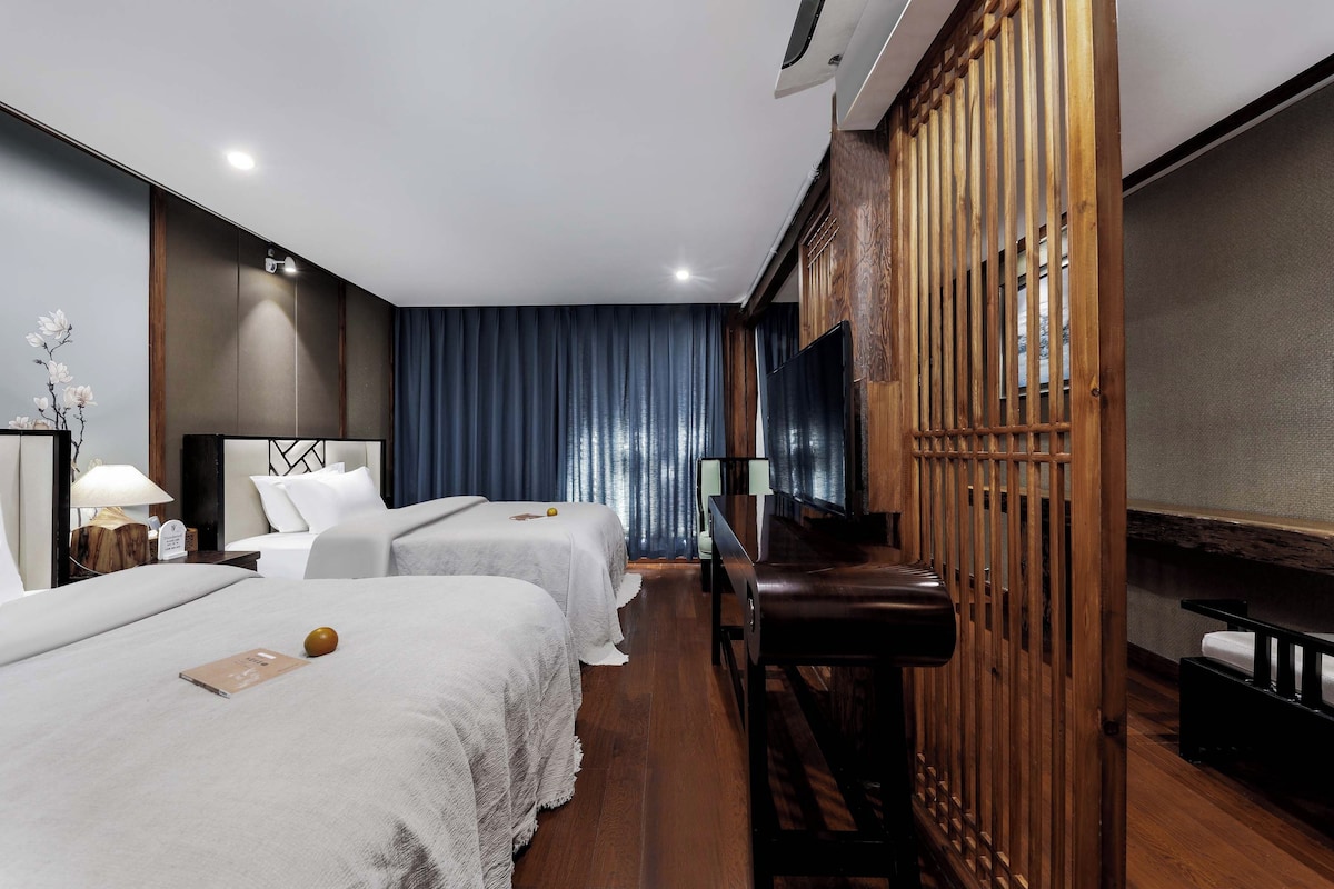 Quarto de hotel com duas camas, uma escrivaninha, uma cadeira, uma televisão e um divisória de madeira.