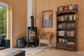 A cozy room with a wood-burning stove, a bookshelf, a wicker chair, and a large window. - Cozy holiday home with spa and sauna (Rødby)