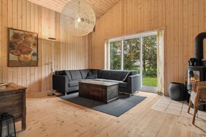 A modern living room with a sectional sofa, a coffee table, and a large window offering a view of greenery. - Cozy holiday home with spa and sauna (Rødby)
