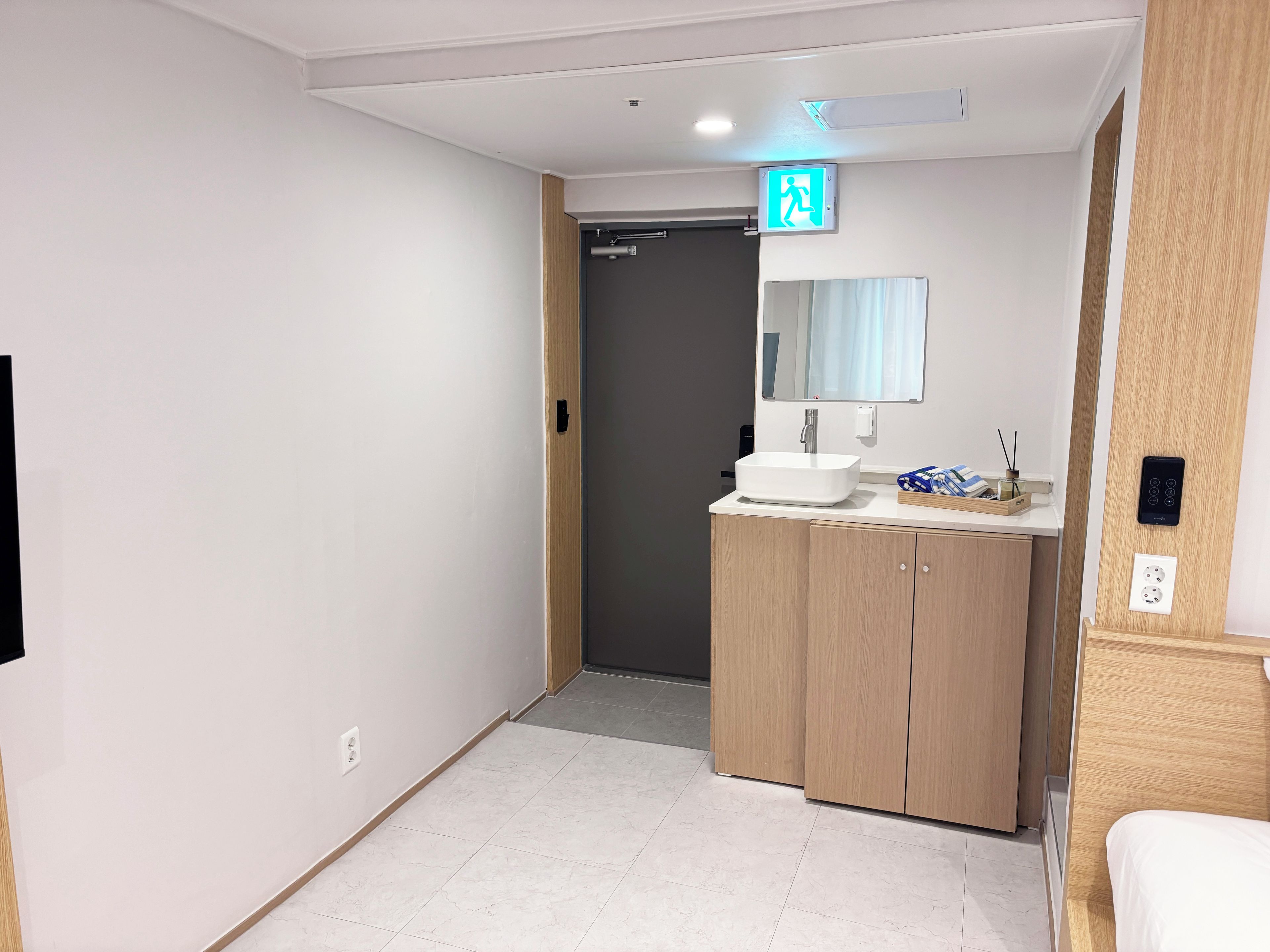 A hotel bathroom with a white sink, wooden cabinets, a mirror, and a visible exit sign.