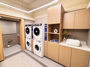 A laundry room with two stacked washing machines, a sink, and a shelf with decorative items. - Monolith Stay Hotel (Incheon)