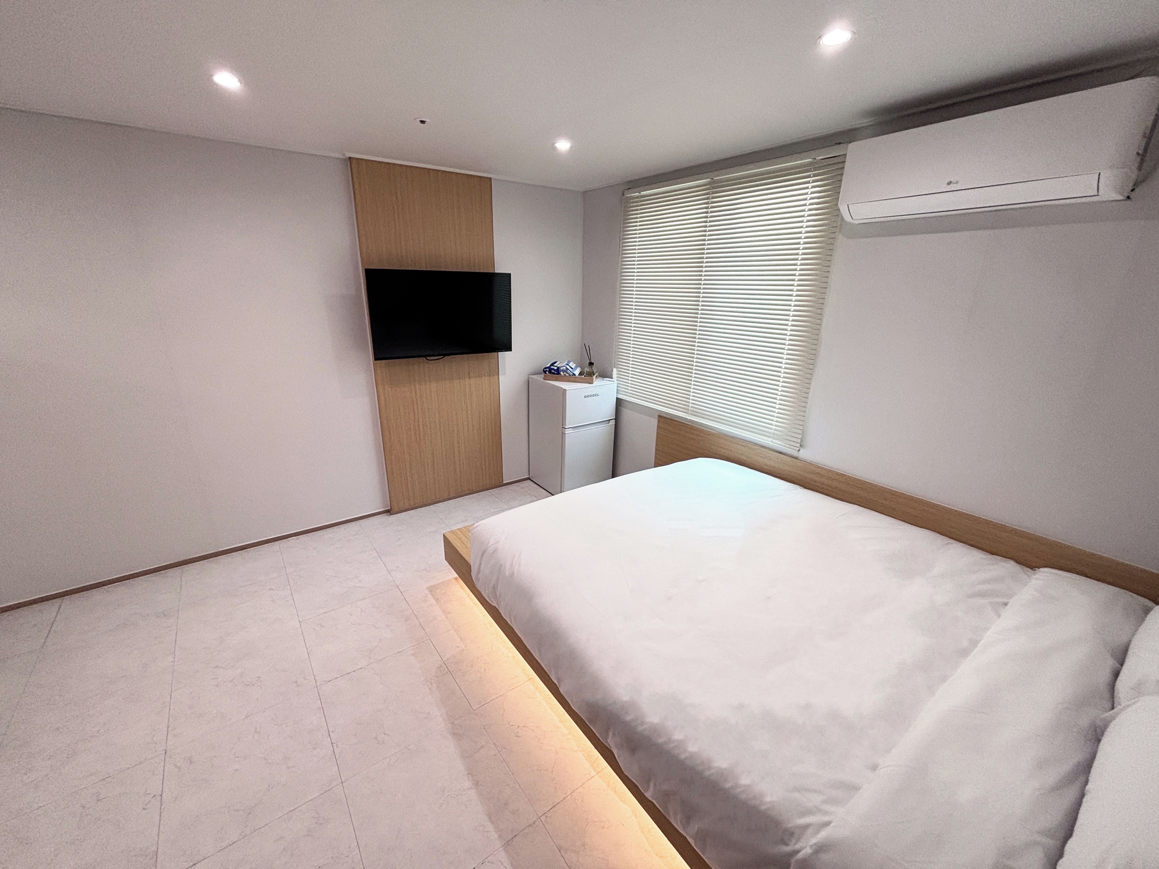 A modern bedroom with a bed, a TV mounted on a wooden panel, a small desk, and a window with blinds.
