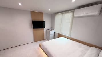 A modern bedroom with a bed, a TV mounted on a wooden panel, a small desk, and a window with blinds.