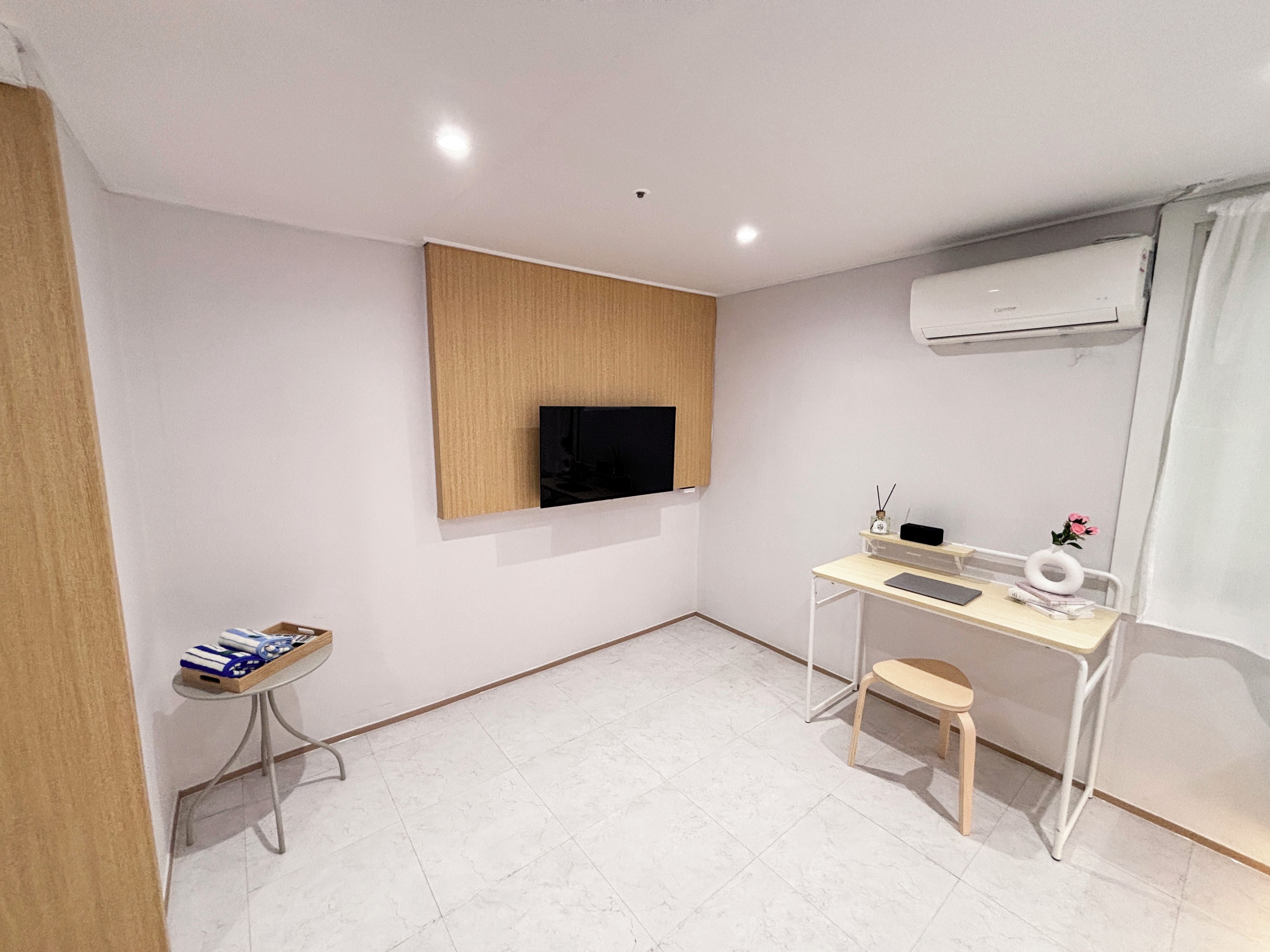 A modern, minimalistic room with a flat-screen TV mounted on the wall, a small desk with a chair, and an air conditioning unit.