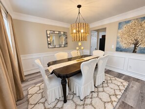A dining room with a dark wooden table, white chairs, a fluffy rug, a chandelier, a mirror, and a painting of a tree. - Spacious Family Retreat Near Stone Mountain Park and Lake Lanier Island (Snellville)