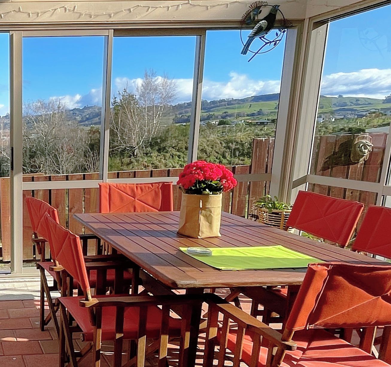Un porche cubierto con una mesa de madera y sillas rojas, un jarrón con flores rojas y vistas a un paisaje montañoso.