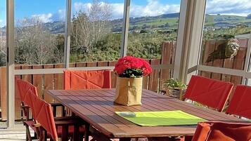 Un porche cubierto con una mesa de madera y sillas rojas, un jarrón con flores rojas y vistas a un paisaje montañoso.
