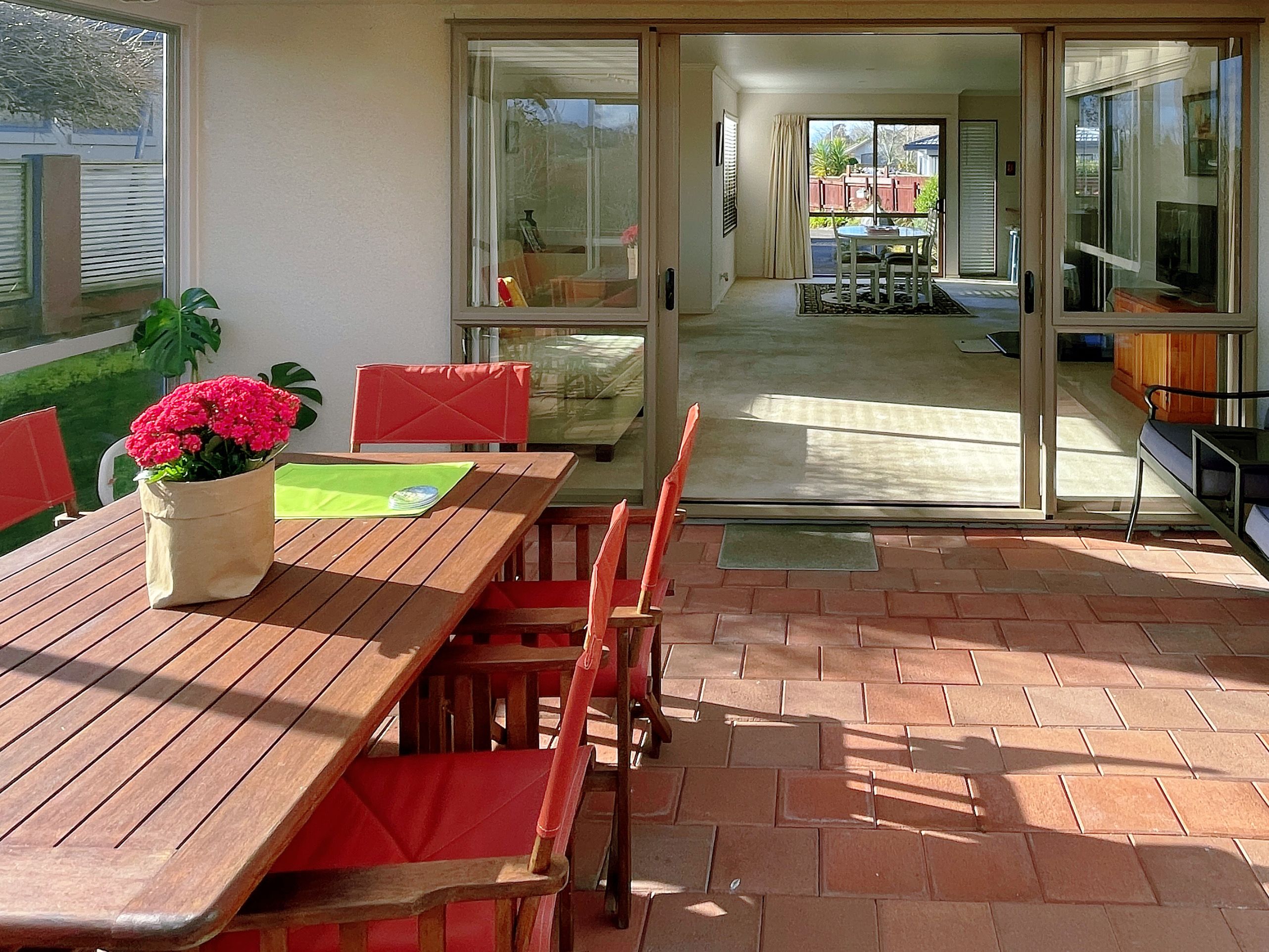 Un patio exterior con mesa y sillas de madera, una planta en maceta y vista al interior de la casa a través de puertas de cristal.