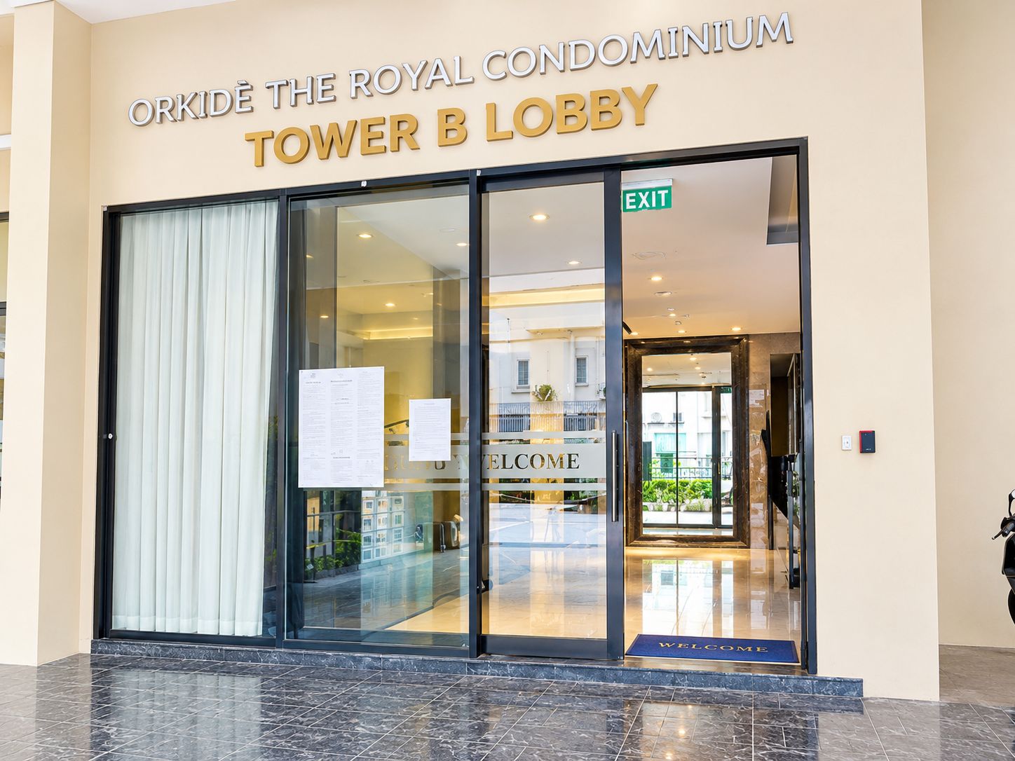 The entrance to the Royal Condominium Tower B Lobby with a glass door, an exit sign, and a welcome mat.