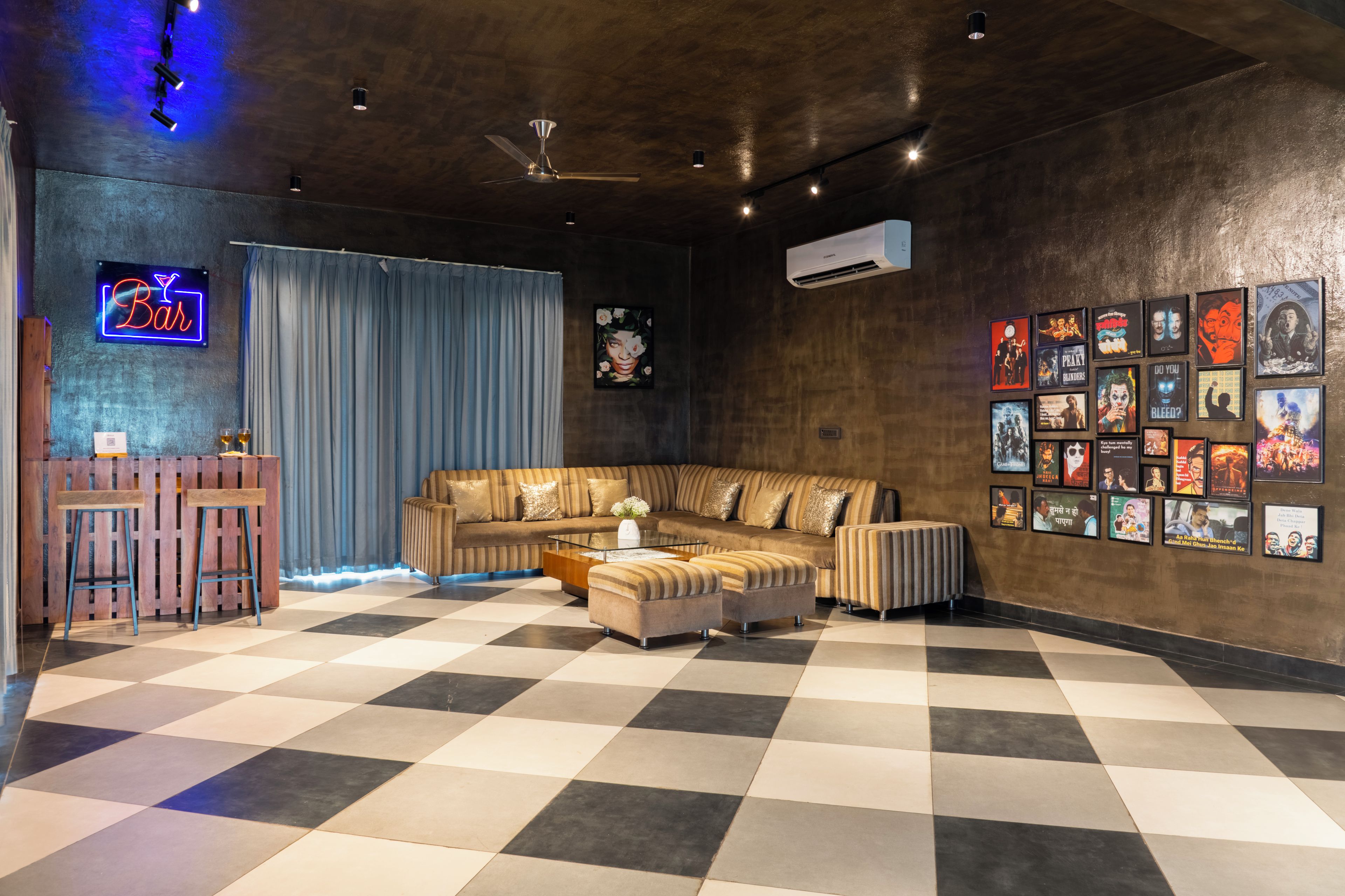 Un salon moderne avec un sol à carreaux, un ensemble de canapés, une table basse, un coin bar avec des tabourets et un mur décoré d’affiches de films.