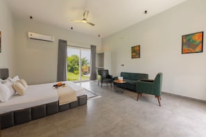 Une chambre d’hôtel moderne équipée d’un lit, d’un canapé, d’une chaise et d’une table à manger. On y trouve une grande fenêtre avec des rideaux, un climatiseur et un ventilateur de plafond.