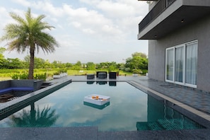 Een modern buitenzwembad met een terras, omringd door groen en een heldere lucht.