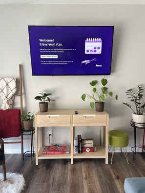 A modern living room with a flat-screen TV displaying a welcome message, a wooden console table with plants, a red throw blanket, and a Crabble board game. - 3 Bedroom, sleeps 8, house centrally located in San Diego (San Diego)