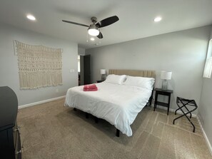 Un dormitorio con cama, ventilador de techo, un macramé en la pared, una mesita de noche con lámpara y una silla.
