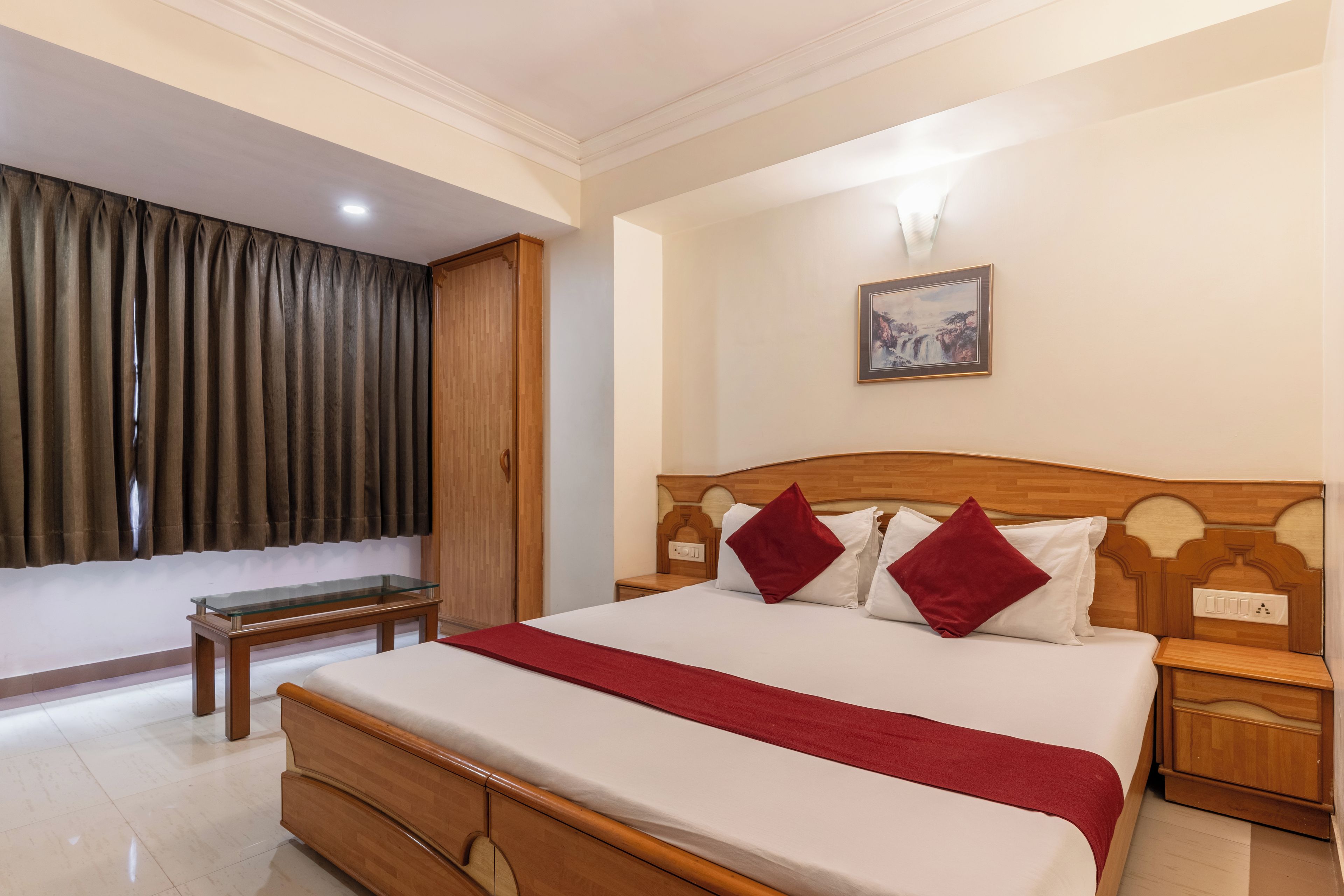 Quarto de hotel com cama de madeira, mesa de cabeceira, luminária e um quadro na parede, além de uma janela com cortinas.