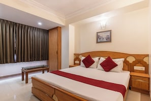 A hotel room with a wooden bed, a bedside table, a nightstand, a wall-mounted painting, and a window with curtains.