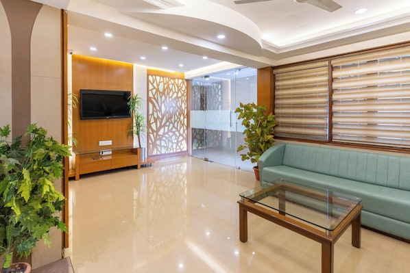 A modern living room with a flat-screen TV, a glass coffee table, a green sofa, and decorative plants.
