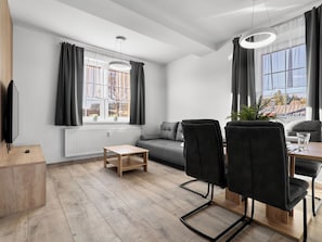 Una sala de estar moderna con un sofá, dos sillas negras, una mesa de centro de madera y una mesa de comedor con sillas. Hay ventanas grandes con cortinas, un radiador y un televisor a la izquierda.