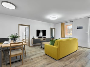 Ein modernes Wohnzimmer mit einem gelben Sofa, einem Essbereich mit einem Holztisch und Stühlen, einem an der Wand montierten Flachbildfernseher und einem Fenster mit Vorhängen.