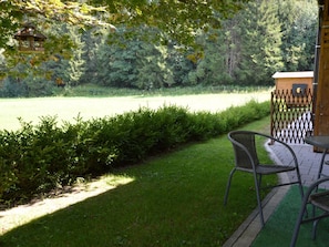 Een terras met uitzicht op een grasveld en bomen. Achtergrond is een houten constructie, mogelijk een tuinhuis of een klein gebouw.
