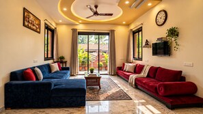 A living room with two sofas, a ceiling fan, a TV, and a large window with curtains. - Valora Stays Shalimar Villa (Lonavala)