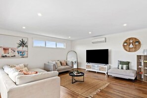 A modern living room with a flat-screen TV, two sofas, a coffee table, and a bookshelf. - Marina Rise - Walk to the Sand (Safety Beach)