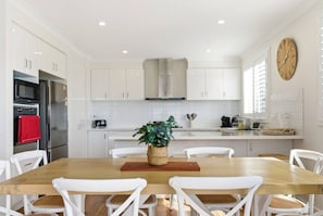 A modern kitchen with white cabinetry, stainless steel appliances, and a wooden dining table with white chairs. - Marina Rise - Walk to the Sand (Safety Beach)