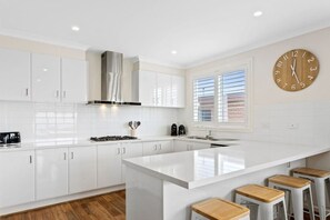 A modern kitchen with white cabinetry, a central island, bar stools, a stove, and a clock on the wall. - Marina Rise - Walk to the Sand (Safety Beach)