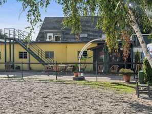 A yellow building with a staircase leading to a balcony, surrounded by a fenced area with outdoor seating and potted plants. - Apartment with a rooftop terrace on a horse farm (Jüchen)