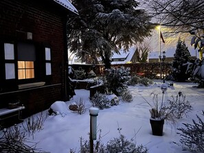 Een bakstenen gebouw met een raam dat een warm interieur toont, omgeven door met sneeuw bedekte planten en een besneeerd landschap.