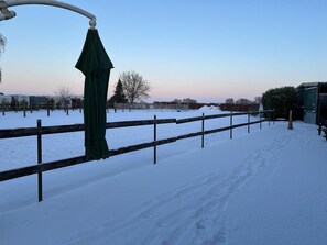 Ein verschneiter Außenbereich mit Holzzaun, einem grünen Sonnenschirm und blauem Himmel.