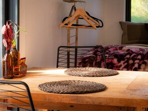 A wooden table with woven placemats, a metal chair, a wooden hanger rack, and a vase with dried plants.