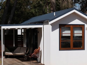 Un petit bâtiment blanc, doté d’un toit sombre, d’une fenêtre à cadre en bois et d’un espace extérieur abrité avec une chaise et une table.
