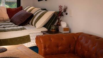 A bedroom with a bed, a brown leather sofa, a bedside table, and a window with a view of greenery.